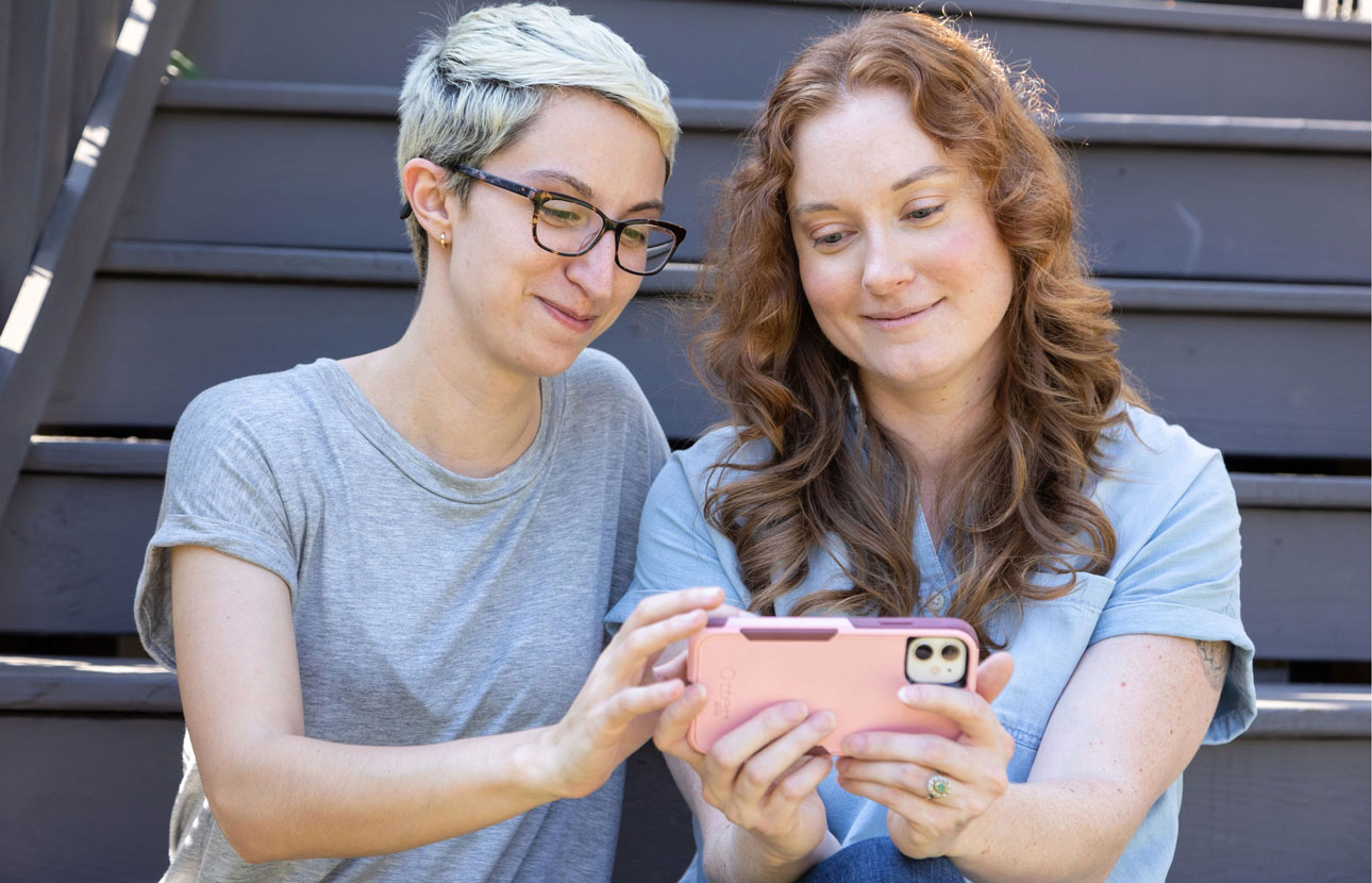 Two women sit on some stairs looking at their smartphone happy about getting Payment Guard coverage on their loan.